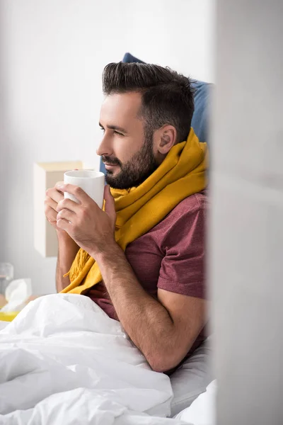 Sorridente malato giovane uomo con sciarpa in possesso di tazza di tè caldo mentre seduto a letto — Foto stock