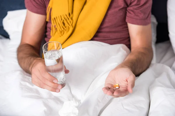 Cropped shot of sick man sitting in bed with glass of water and pills — Stock Photo