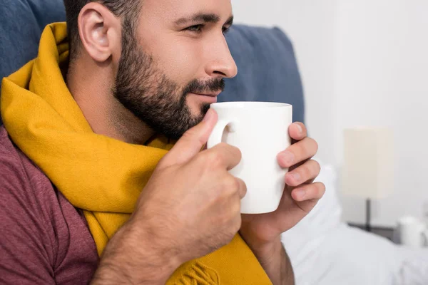 Nahaufnahme Porträt eines lächelnden kranken jungen Mannes mit Schal, der eine Tasse heißen Tee im Bett hält — Stockfoto
