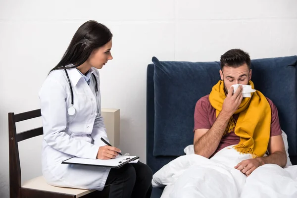 Médico escribiendo diagnóstico en portapapeles mientras el paciente estornuda y se sienta en la cama - foto de stock