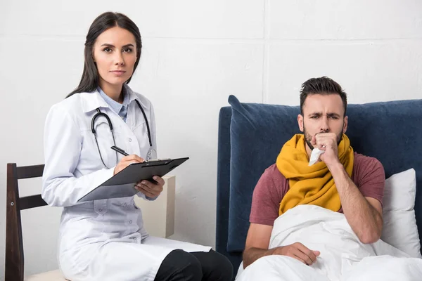 Médico escribiendo diagnóstico en portapapeles mientras el paciente tiene tos y sentado en la cama - foto de stock