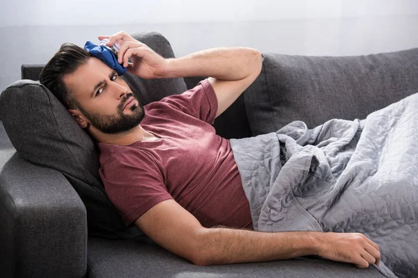 Kranker junger Mann liegt auf Couch und hält Eisbeutel auf der Stirn und blickt in die Kamera — Stockfoto