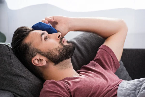 Close-up portrait of sick young man lying on couch and holding ice pack on forehead — Stock Photo