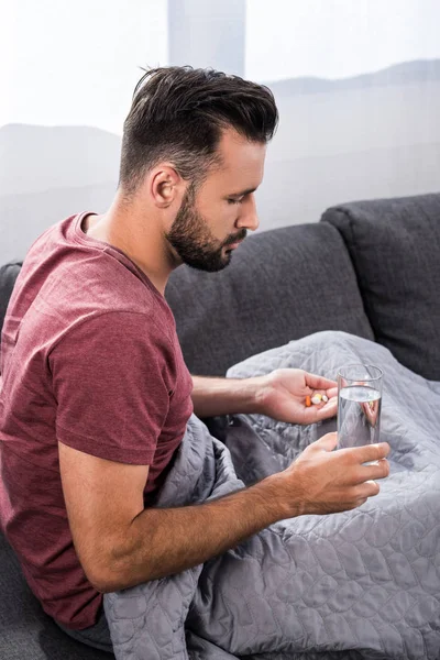 Хворий молодий чоловік сидить на дивані зі склянкою води і таблеток — стокове фото