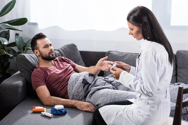 Doctora sosteniendo termómetro eléctrico mientras su paciente acostada en el sofá y hablando con ella - foto de stock