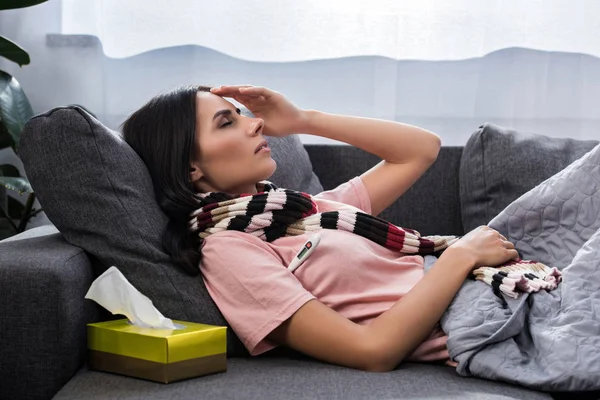 Mujer joven enferma que mide la temperatura con termómetro eléctrico en el sofá — Stock Photo