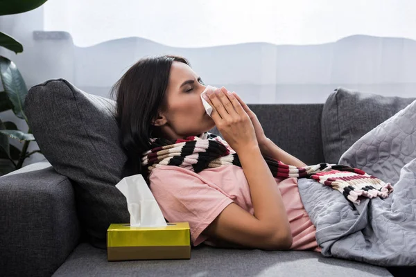 Diseased young woman sneezing into paper napkin on sofa — Stock Photo