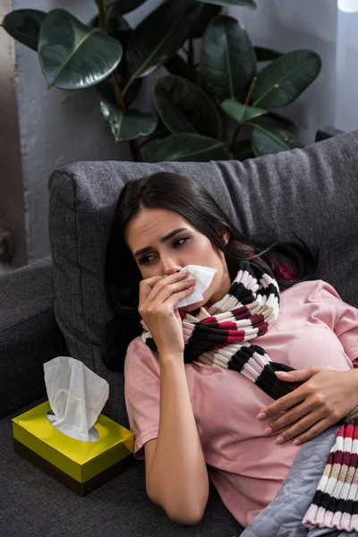 Diseased young woman sneezing into paper napkin on couch — Stock Photo