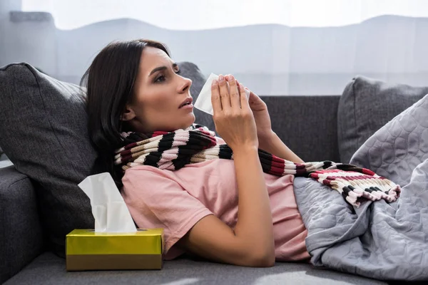 Sick young woman sneezing into paper napkin on couch — Stock Photo