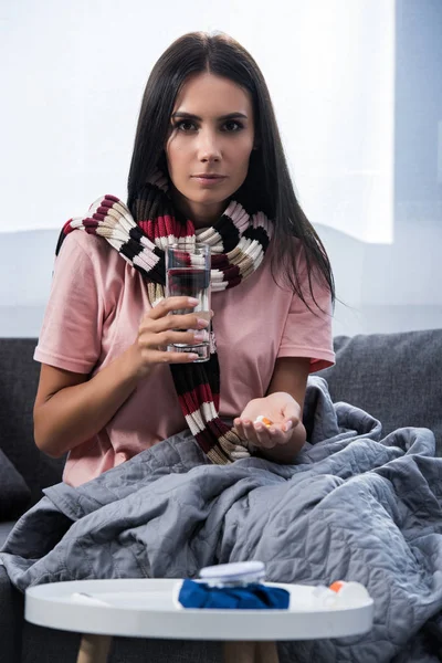 Kranke junge Frau mit Glas Wasser und Pillen auf Couch sitzend und in die Kamera blickend — Stockfoto