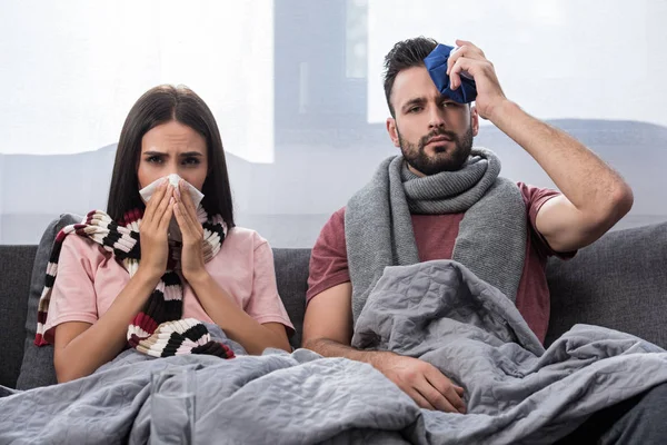 Enferma joven pareja con servilleta y hielo paquete sentado juntos en el sofá y mirando a la cámara - foto de stock