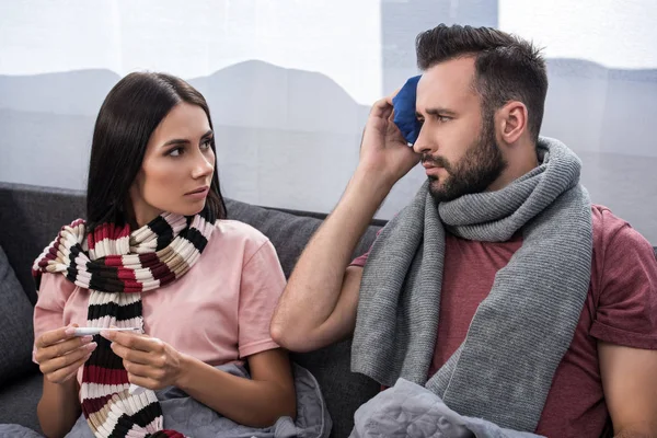 Sick young couple with electric thermometer and ice pack sitting together on couch — Stock Photo