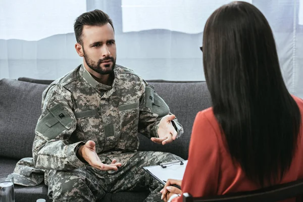 Sad soldier with ptsd talking at psychiatrist and gesturing while sitting on couch during therapy session — Stock Photo