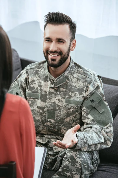 Soldado sonriente hablando al psiquiatra y haciendo gestos mientras está sentado en el sofá durante la sesión de terapia - foto de stock
