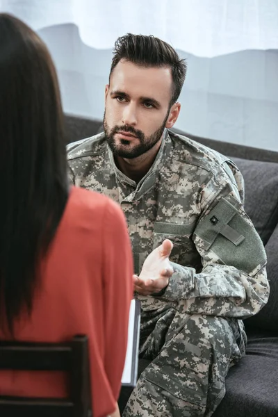 Soldado deprimido hablando en psiquiatra y haciendo gestos mientras está sentado en el sofá durante la sesión de terapia - foto de stock