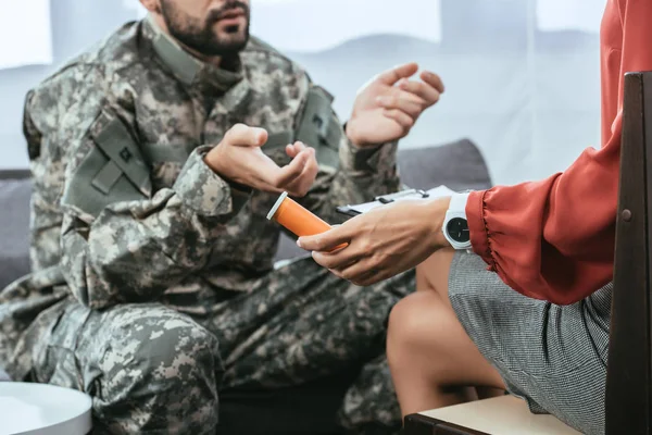 Recortado tiro de soldado en uniforme con ptsd hablando con psiquiatra en sesión de terapia mientras ella sosteniendo frasco de píldoras - foto de stock