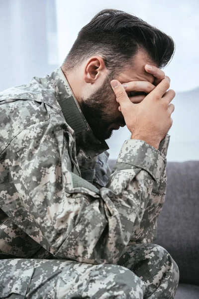 Vista lateral del hombre deprimido del ejército en uniforme militar con trastorno de estrés postraumático sosteniendo su cabeza mientras está sentado en el sofá - foto de stock