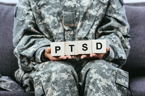 Cropped shot of female soldier in military uniform sitting on sofa and holding wooden cubes with PTSD sign — Stock Photo