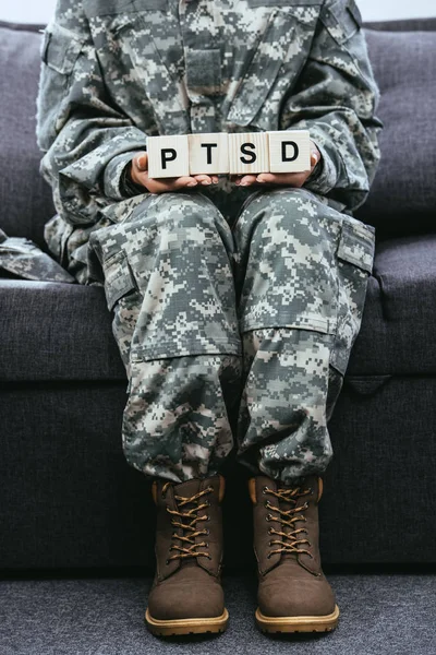 Tiro recortado de soldado fêmea em uniforme militar sentado no sofá e segurando cubos de madeira com sinal PTSD — Fotografia de Stock