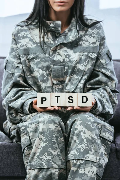 Cropped shot of young female soldier in military uniform sitting on couch and holding wooden cubes with PTSD sign — Stock Photo