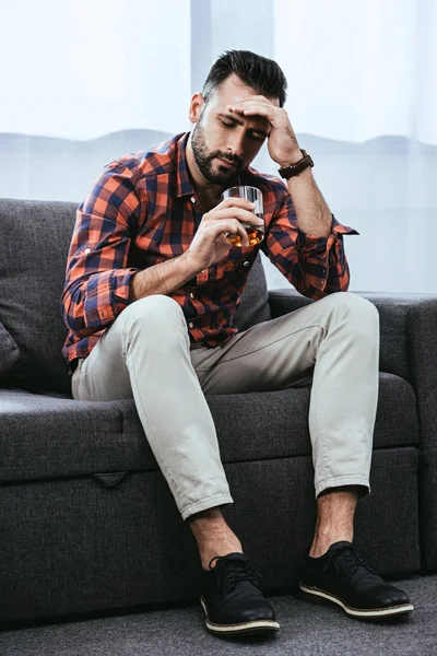 Depressed young man with glass of whiskey sitting on couch at home alone — Stock Photo