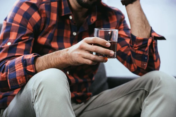 Cropped shot of young man holding glass of whiskey while sitting on couch — Stock Photo