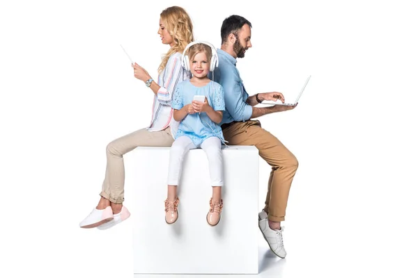 Family using digital devices and sitting on white cube isolated on white — Stock Photo