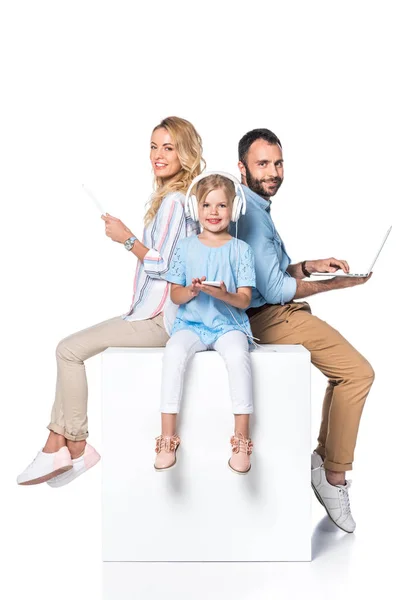 Familia usando aparatos digitales y sentado en cubo blanco aislado en blanco - foto de stock