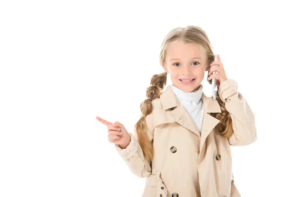 Niño sonriente en abrigo beige hablando en el teléfono inteligente y mostrando algo, aislado en blanco - foto de stock