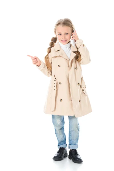 Adorable enfant en manteau beige parlant sur smartphone et montrant quelque chose, isolé sur blanc — Photo de stock