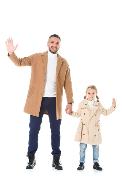 Elegante padre e hija en abrigos beige tomados de la mano y saludando aislado en blanco - foto de stock