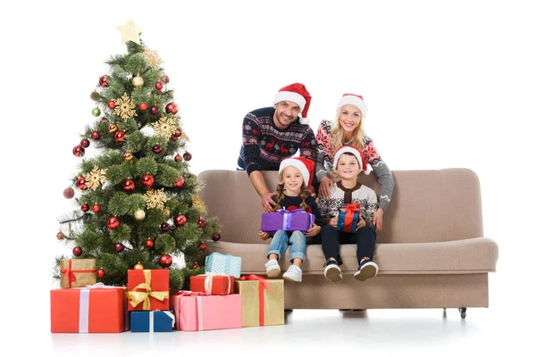 Familia sonriente con niños en sombreros de santa sentado en el sofá cerca del árbol de Navidad con cajas de regalo, aislado en blanco - foto de stock