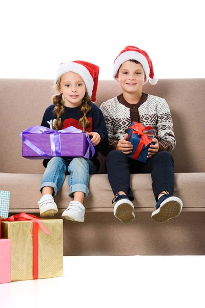 Feliz hermano y hermana en sombrero de santa sentado en sofá con regalos de Navidad, aislado en blanco - foto de stock