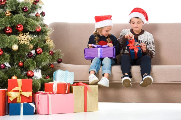 Frère et soeur dans le chapeau santa tenant des cadeaux et assis sur le canapé près de l'arbre de Noël, isolé sur blanc — Photo de stock