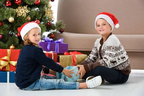 Hermanos felices en sombrero de santa sentado cerca del árbol de Navidad con regalos - foto de stock