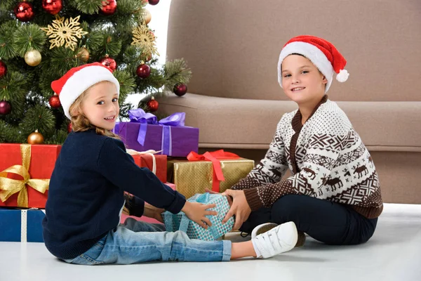 Irmãos felizes em santa chapéu sentado perto da árvore de natal com presentes — Fotografia de Stock