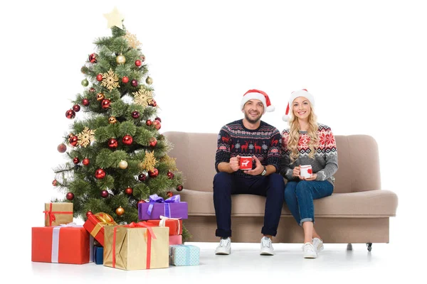 Casal feliz com xícaras de café e sentado no sofá perto da árvore de natal com presentes, isolado em branco — Fotografia de Stock