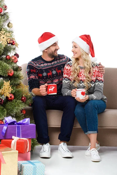 Beautiful couple in santa hats holding cups of coffee and sitting on sofa near christmas tree with gifts, isolated on white — Stock Photo