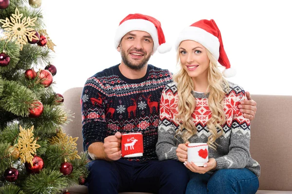 Bella coppia in cappelli di Babbo Natale in possesso di tazze di caffè e seduto sul divano vicino all'albero di Natale, isolato su bianco — Foto stock