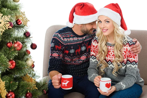 Glückliches Paar in Weihnachtsmannmützen, Kaffeetassen in der Hand und am Weihnachtsbaum sitzend, isoliert auf weiß — Stockfoto