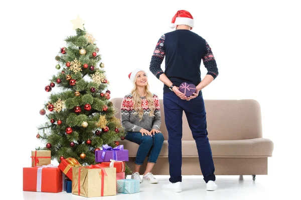 Frau sieht Mann an, der herzförmiges Geschenk versteckt und in der Nähe des Weihnachtsbaums steht, isoliert auf weiß — Stockfoto