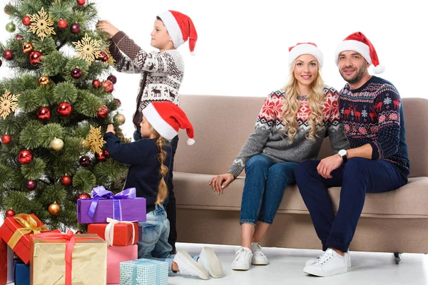 Children decorating christmas tree while happy parents sitting on sofa near, isolated on white — Stock Photo