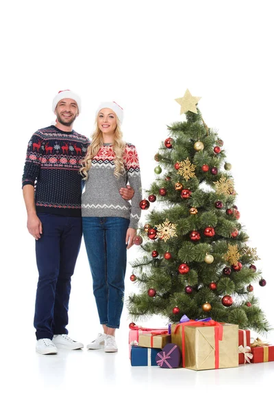 Hermosa pareja feliz en sombreros de santa de pie cerca del árbol de Navidad con regalos, aislado en blanco - foto de stock