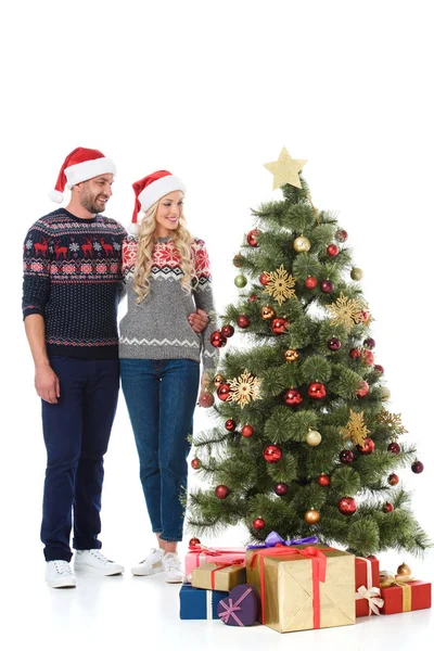 Smiling couple in santa hats looking at christmas tree with presents, isolated on white — Stock Photo