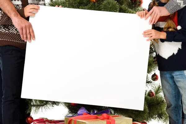 Ausgeschnittene Ansicht von Familie, die leeres Brett gegen Weihnachtsbaum hält — Stockfoto