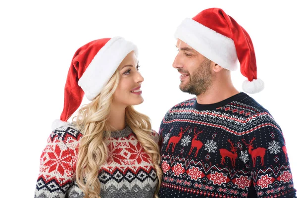 Pareja sonriente en suéteres de Navidad y sombreros de Santa Claus mirándose, aislados en blanco - foto de stock