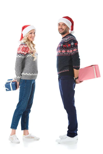 Casal em camisolas de Natal e chapéus de Papai Noel escondendo caixas de presente, isolado em branco — Fotografia de Stock