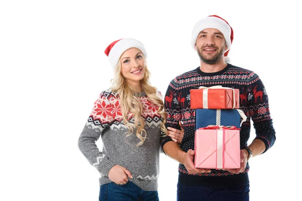 Smiling couple in christmas sweaters and santa hats holding gifts, isolated on white — Stock Photo