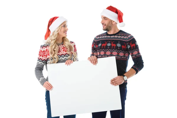 Couple en chandails de Noël et chapeaux de Père Noël tenant une pancarte vide, isolé sur blanc — Photo de stock