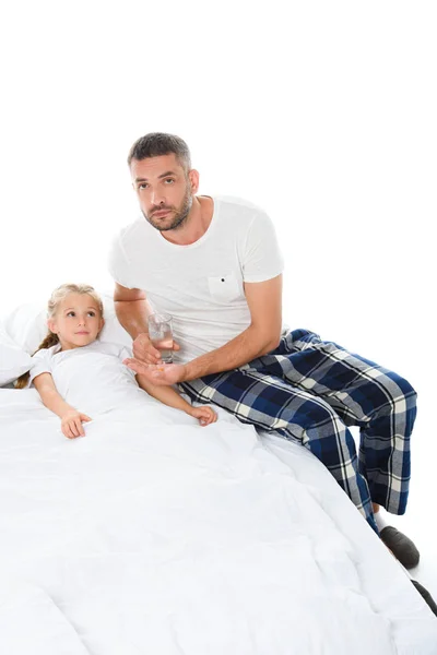 Padre adulto dando pastillas y vaso de agua a la hija enferma acostada en la cama, aislada en blanco - foto de stock